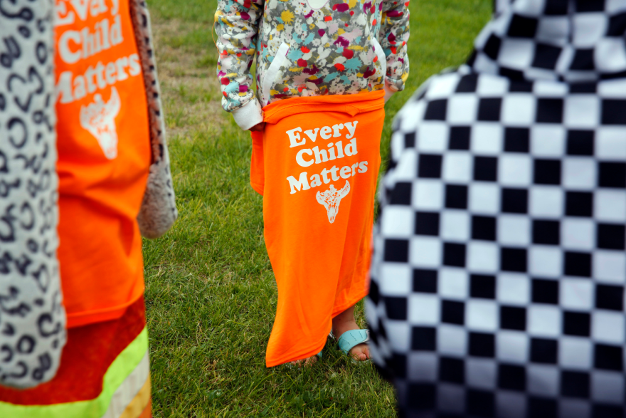 A child, visible only from the waist down, holds an orange shirt that says Every Child Matters and has an animal head on it, possibly a bull. Another child in the foreground, visible from the chest down, is wearing one of those shirts.