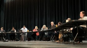 Nine people can be seen sitting behind tables on a stage. On the tables, sit laptops and microphones for each person. 