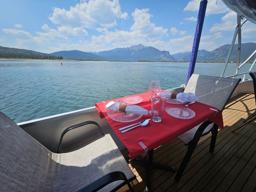 a table set for two on the deck of a boat has a red tablecloth, two beige and brown chairs. It's overlooking the water of Lake Dillon with mountains in the background