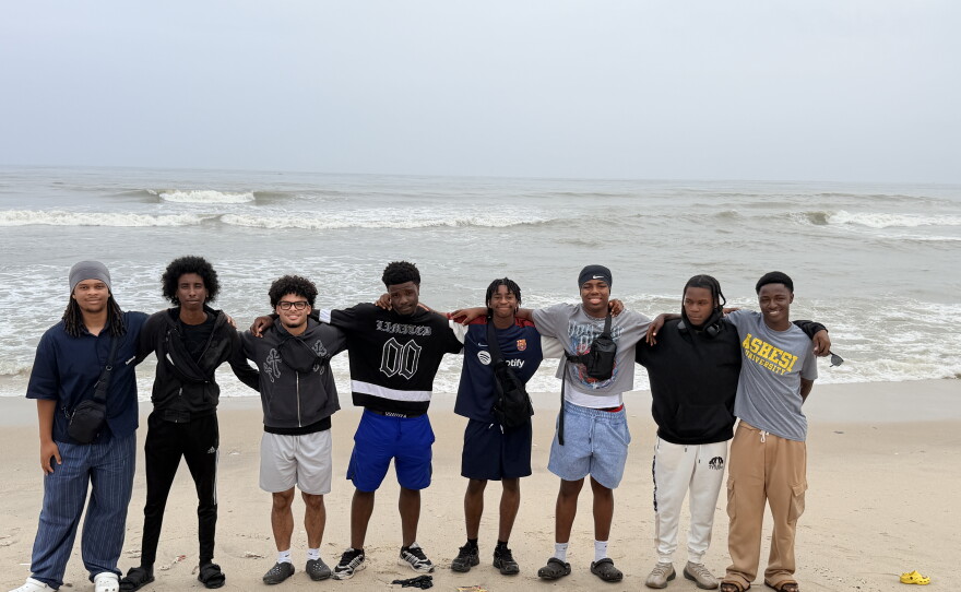 A group of young men stand in a line with their arms around each other in front of an ocean.