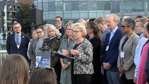 A group of civic leaders stands around former Washington Gov. Chris Gregoire on a rooftop. Gregoire is speaking at a podium into several microphones. A sign on the podium says Partnership for a Competitive Puget Sound. 