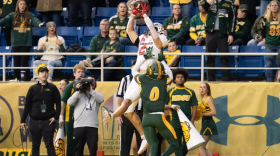 A receiver jumping up to catch a football against a defender reaching up to him and fans, cheerleaders, staff and a camera operator watching in the background. 
