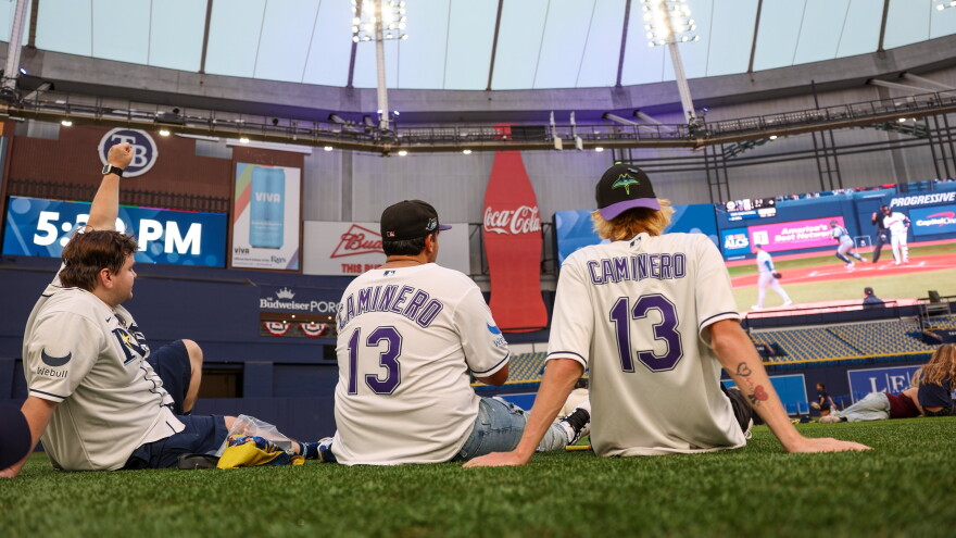 3 men in rays baseball jerseys sit on an astroturf field inside a stadium and watch a baseball game on a huge ballpark videoboard