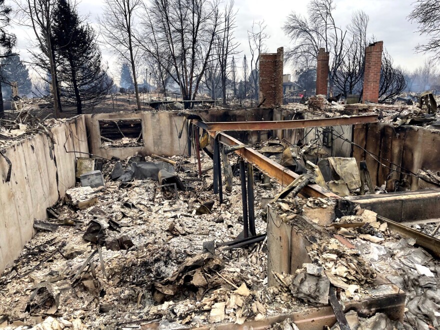 The burnt remains of a home lie charred and ashy inside the burnt white frame of a house with trees in the background.