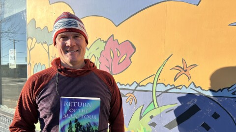 A man wearing a red an maroon sweater holding a book in front of mural or lakes and plants.