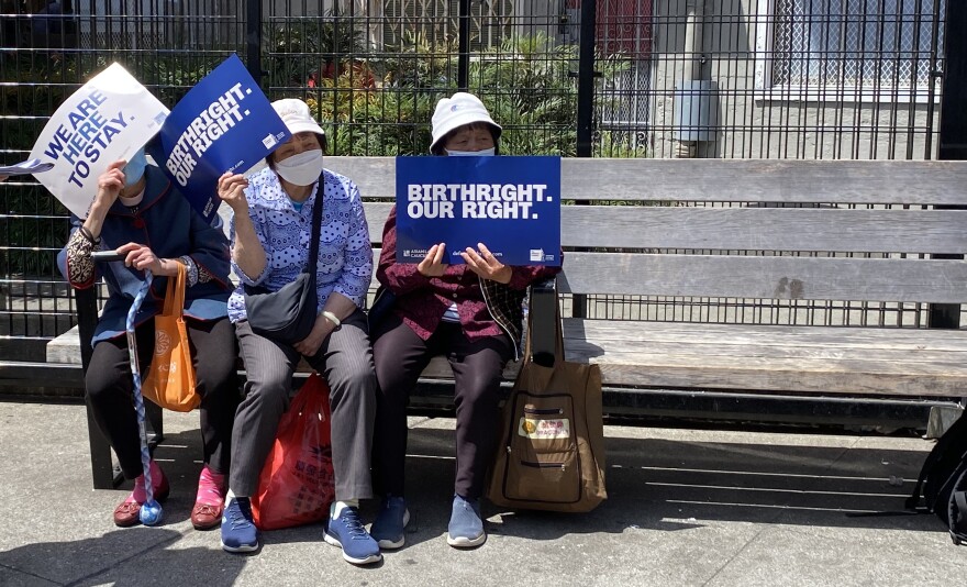Three elder Asian women sit on a bench in San Francisco Chinatown, holding signs that read "BIRTHRIGHT. OUR RIGHT." and "WE ARE HERE TO STAY".