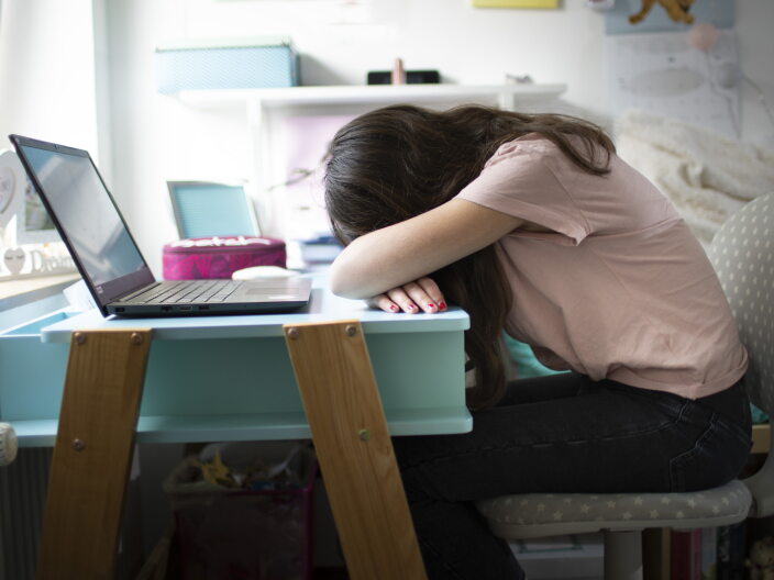 A girl sits at a desk in front of a laptop, resting her head on her arms.