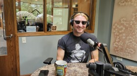 A person sitting in a radio studio with a microphone near their mouth. They have on black sunglasses.