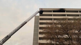 Fire trucks with extended ladder respond to a fire on the upper floor of a multi-story building, with black smoke visible from the windows. Trees and emergency lights are also visible in the scene.