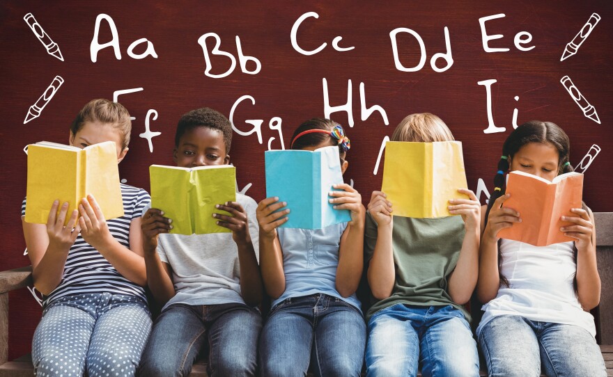 Various children are sitting in a row, books held in front of their faces