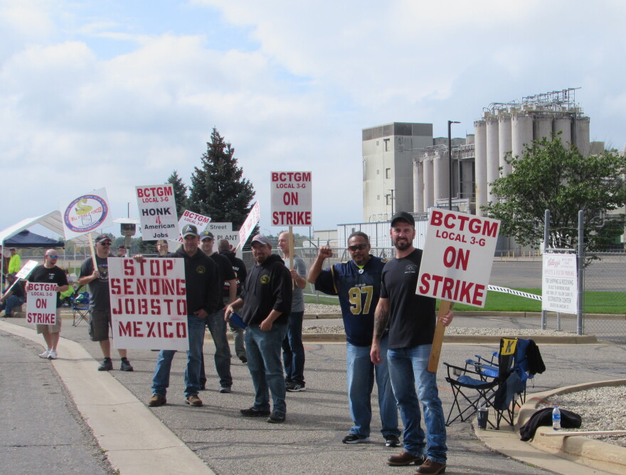 a group of workers stands along a road with the grain silos of the plant in the background. The picketers carry signs with red lettering.
