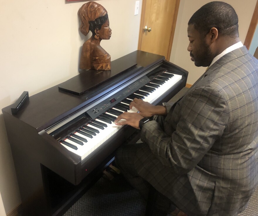 A Black man with his back to the camera in a gray and tan checkered suit sits at a keyboard and plays a song 