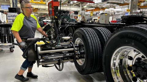 A woman wearing black gloves and glasses is using a machine to help attach wheels to a truck.