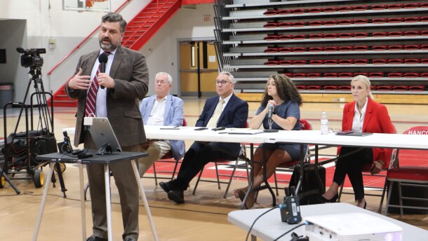 A man in a suit and tie speaks into a microphone in a school gymnasium, with members of the Kentucky Public Service Commission seated at a table behind him.