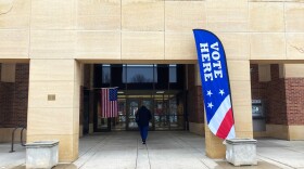 a woman walks into a brick building. there is an American flag and a sign that says VOTE HERE
