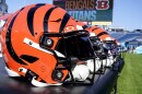 a row of orange helmets with black stripes sits on a black table on the sidelines of a green football field