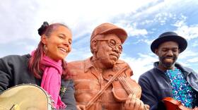 This image was captured during a reunion of Rhiannon Giddens and Justin Robinson, funding members of The Carolina Chocolate Drops in Mebane, North Carolina. The image includes part of the memorial to Joe and Odell Thompson known as "Knee to Knee" Joe & Odell Thomson, 2019 by sculptor Brad Spencer. The mason who molded the bricks was Tommy Whitmore. In the photograph, Giddens and Robinson appear next to fiddler Joe while banjo player Odell Thomson was cropped out. The Photograph was captured on November 2, 2021 during the filming of the documentary "Black Fiddlers".