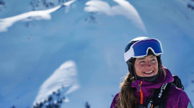 A woman wearing a ski helmet, goggles, jacket and backpack smiles. Behind her is a massive snowy peak dazzled by sunlight.