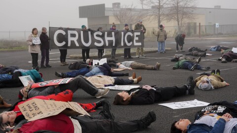 Demonstrators hold a sign reading "crush ice" with people laying on the ground of a parking lot in the foreground. 