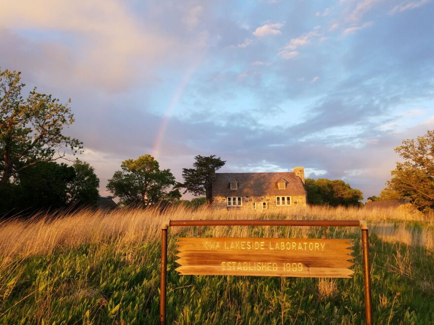 Beautiful photo of prairie with an old stone cabin. There is a faint rainbow overhead. In the foreground there's a sign that says Iowa Lakeside Laboratory established 1909.