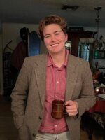 KERA News reporter Kailey Broussard poses in a green tweed suit, red button-down shirt holding a brown coffee mug.