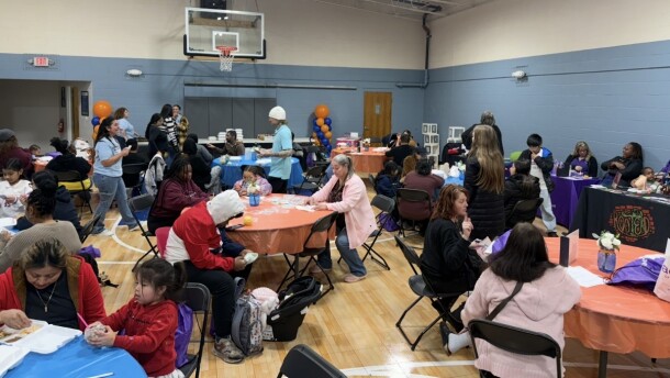 Tables with orange and blue plastic tablecloths are set up around a small community center gym. People of all ages sit around the tables, drawing and talking. There are balloons and baby decorations around the room, and the walls are lined with people at long tables presenting information.