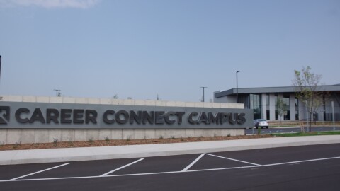 A large, shiny gray sign reads "Career Connect Campus" in black text. A large and long building looms behind the sign, with rows of glass windows and pale gray metal siding lining the outside of the facility. 