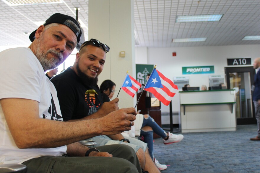 A father and son holding Puerto Rican flags smile at the Frontier Airlines gate.