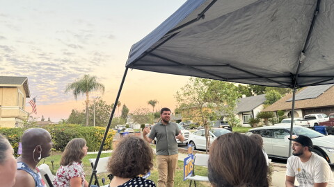 A man talks facing the camera on a driveway in a neighborhood while people watch under a tent 