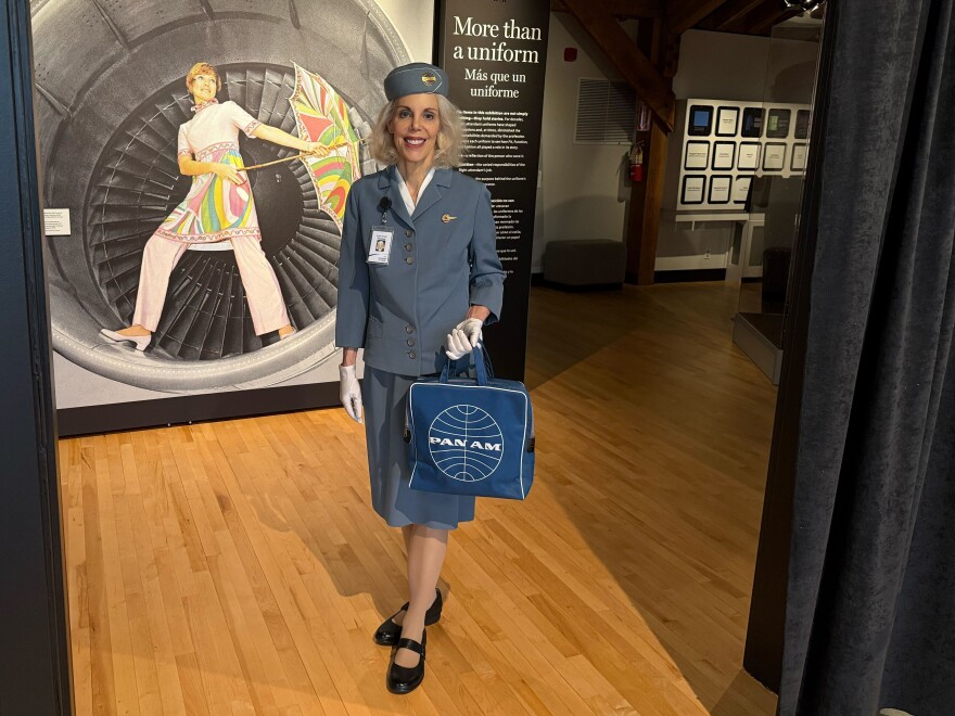A woman wearing a blue vintage outfit with white gloves and a blue Pan Am bag stands at the entrance to a dimly lit room. Behind her is a photo of a woman wearing a pink dress and holding a colorful umbrella while standing in the well of a plane's engine. 