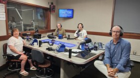 Four people wearing headphones sit at a table in a radio talk studio: a woman front left has short brown hair and is wearing a floral patterned white t-shirt, black shorts and white sandals; a man front right has short grey hair and is wearing eyeglasses, a light blue button-down shirt and white pants; a man back left has short grey hair and is wearing eyeglasses and a beige button-down shirt; a man back right has a long dark ponytail and a dark goatee and is wearing a white t-shirt with a black picture on the chest.