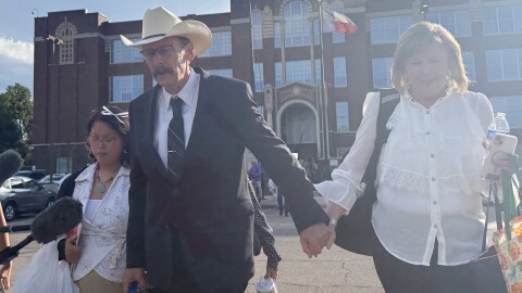 A man with a mustache wearing a suit, glasses and a cowboy hat walks holding hands with a woman in a white blouse. 