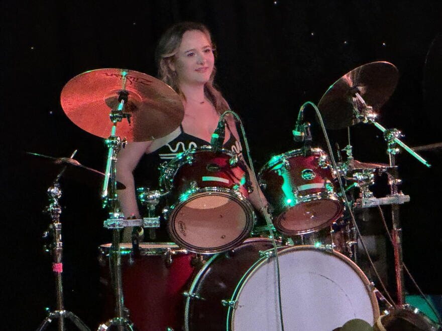A young woman smiling while playing a drum set on a stage.