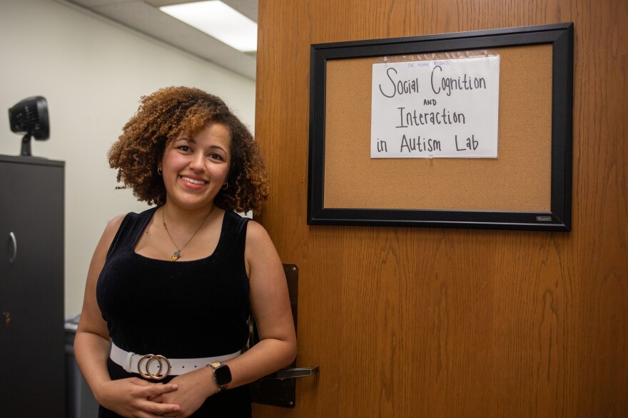 A woman poses for a portrait in front of a sign that reads "Social Cognition and Interaction in Autism Lab".