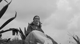 A black and white photo of a woman in braids sitting on a desert terrain. 