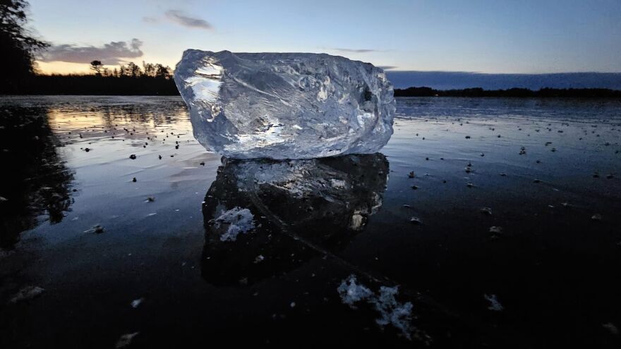 A large of ice sits on thick, clear, smooth lake ice at sunset.