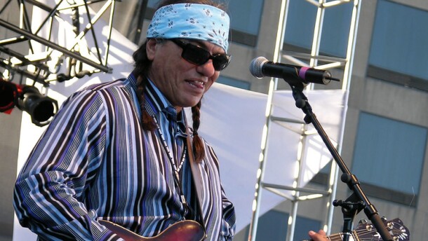 A man in a blue bandana and sunglasses plays the electric guitar in front of a mic stand