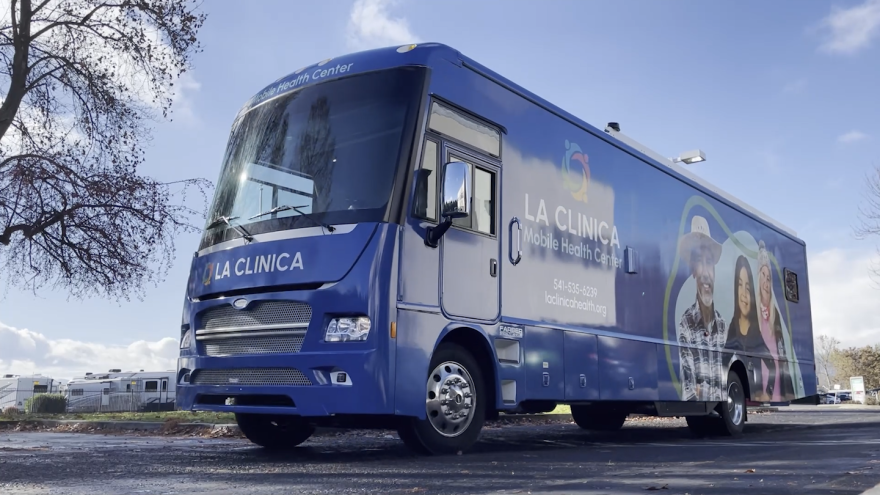 A large blue bus is parked in a parking lot. It reads "La Clinica Mobile Health Center."