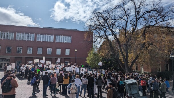A crowd of students gather as students facing the crowd hold signs at a student union protest in front of a brick building under a blue sky.