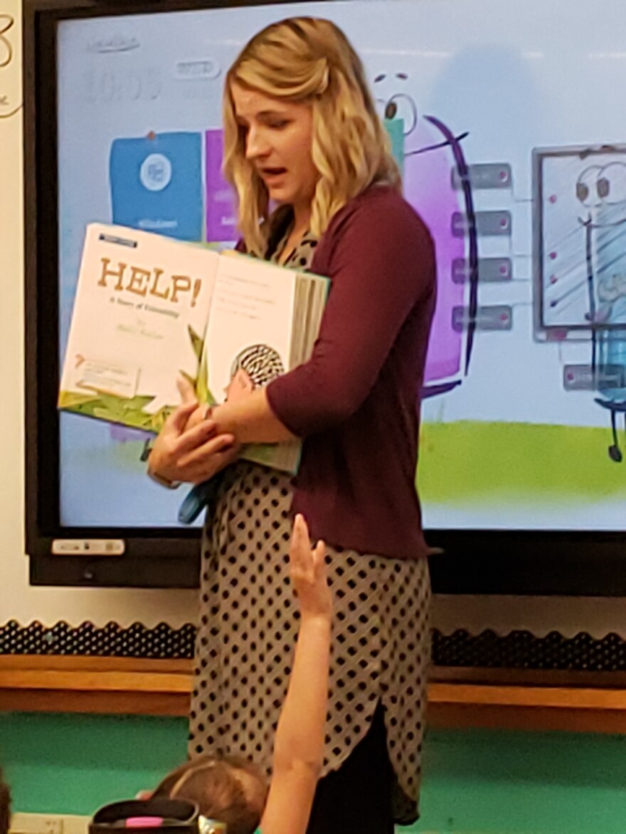 A woman in a red sweater and dress stands holding a book in front a classroom of second grade students.