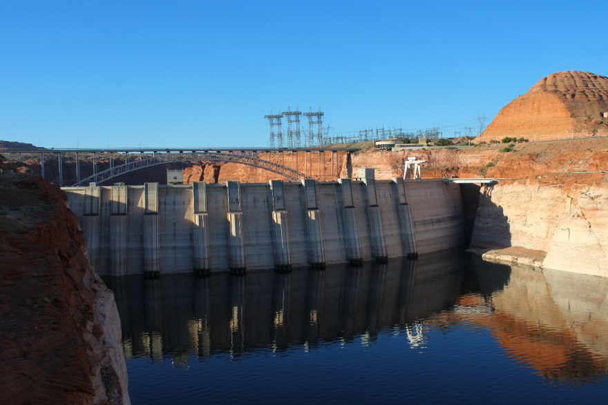 A dam holds back a river near red rocky outcroppings.