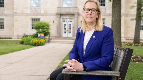 A white woman with shoulder length blonde hair, glasses, a pearl necklace and blue blazer sits on a bench looking somberly at the camera with her hands clasped 