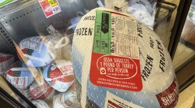 A netted bag holding a 7.42 lb frozen turkey is suspended in a grocery freezer door over several dozen other frozen turkeys on sale. The label on the nearer turkey says, USDA 1 pound of turkey per person.
