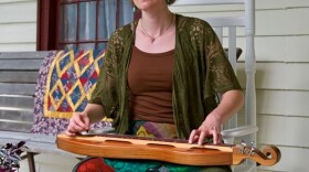 Roxanne McDaniel is sitting on a rocking chair on a porch with a dulcimer in her lap.