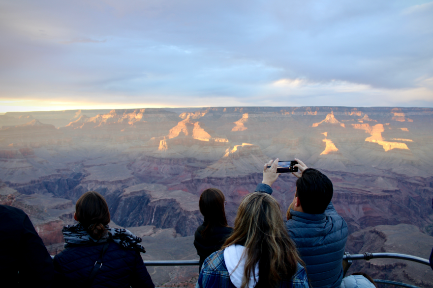Several people standing and taking pictures near a railing overlooking part of the Grand Canyon. The rocky formations go out far into the distance, with the drop-off into the canyon itself visible relatively close by.