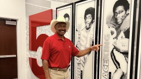 A man in a red University of Georgia polo and white cowboy hat stands in a hallway beside a large red “5,” smiling and pointing at a framed football player portraits on the wall.