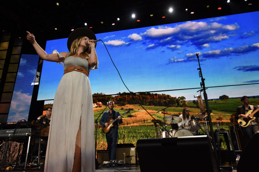 Margo Price sings in front of a backdrop featuring bright blue skies and rolling fields.png