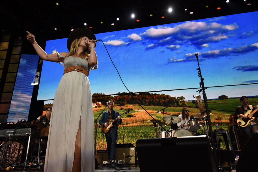 Margo Price sings in front of a backdrop featuring  bright blue skies and rolling fields.png