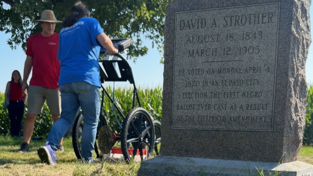 a memorial gravestone is in the foreground. In the background, a volunteering pushes an underground radar device while a man in a red t shirt watches