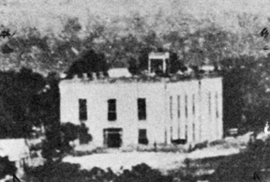  An old, low quality photograph of a two story white stone building. 